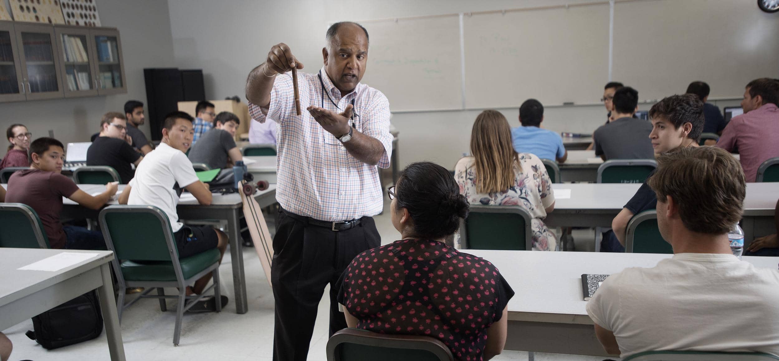 A dark-skinned middle-aged man teaching a classroom full of students.