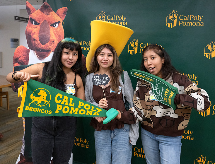 Three girls posing in front of a Cal Poly Pomona branded backdrop, with the outer two pointing towards the center girl