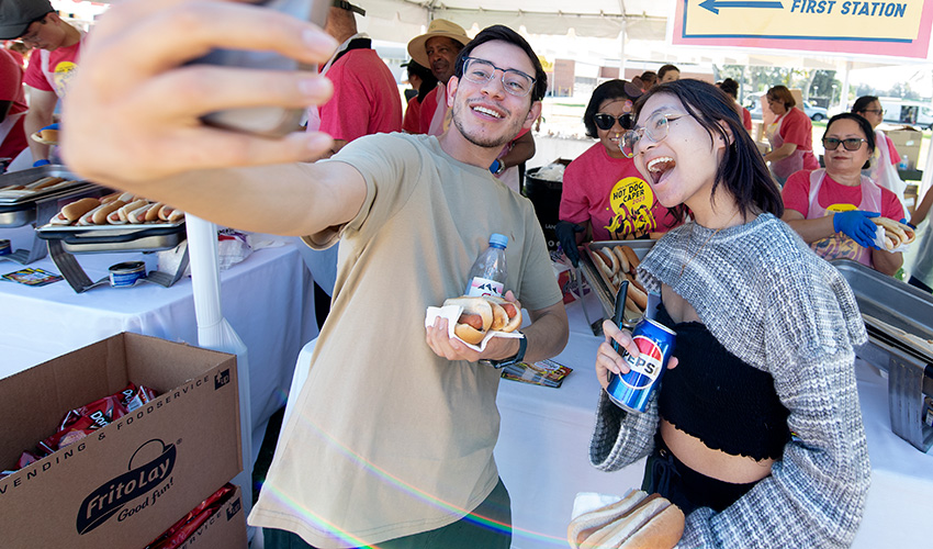 A male and female student take a photo at a Hot Dog Caper tent. 