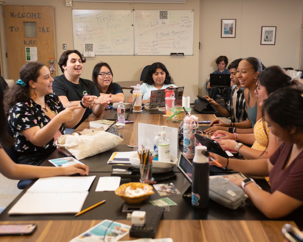 Students participate in a club meeting, smiling and working together at a large table covered with notebooks, snacks, and devices.