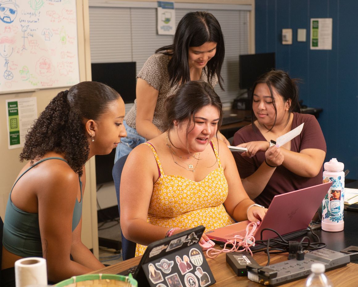 A group of students gathers around a laptop, discussing and working together on a collaborative project in a study space.