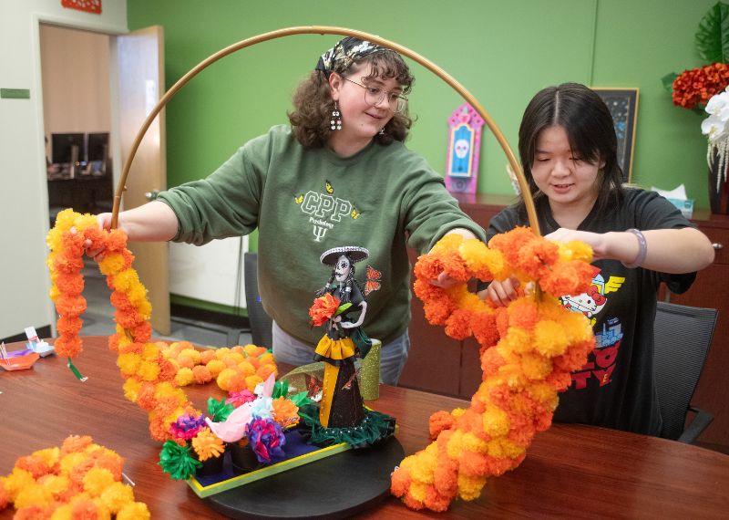 Two students collaborate on a Día de los Muertos altar decoration, arranging marigold flowers and colorful paper crafts.