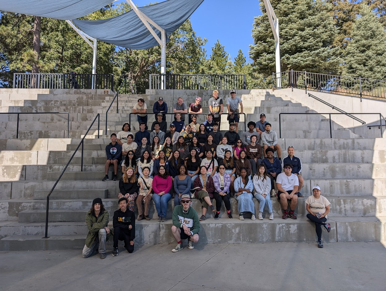 Large group of students sitting on outdoor concrete bleachers under a shaded canopy.
