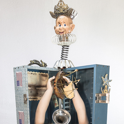 Gina M.  Not A Drill from the Toy Box Kids Series, 2018  assemblage, blue box, doll parts, spring, gold table, horn, cabinet hardware  30 x 18 x 12”  Courtesy of the artist