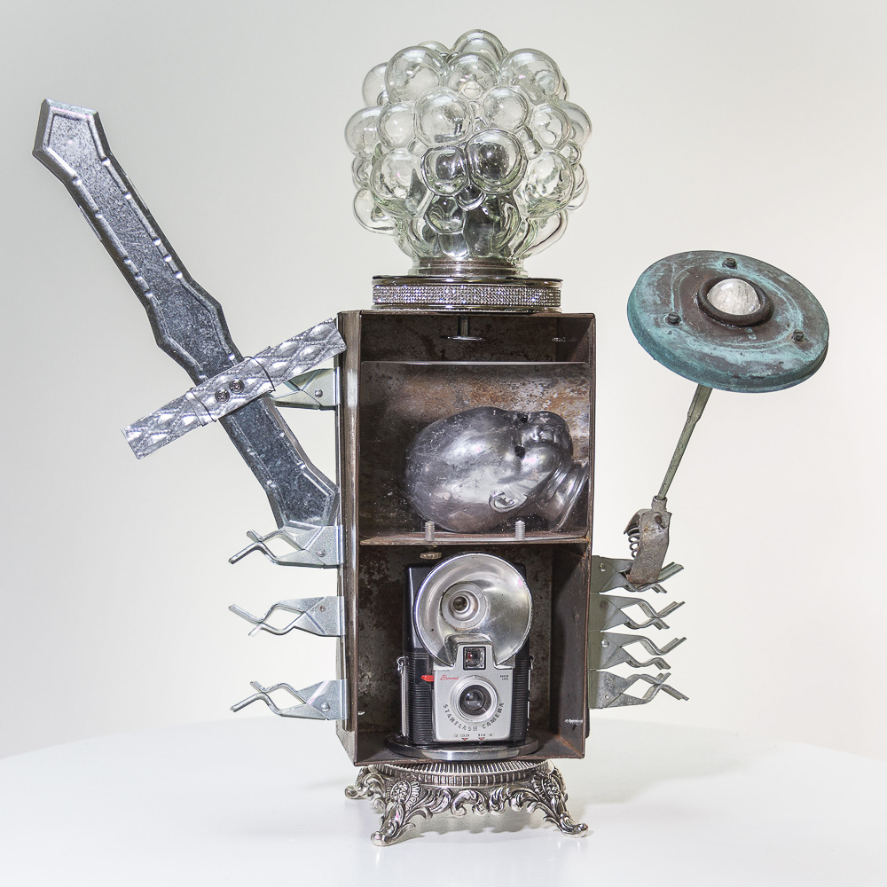 Gina M.  Knight of the Truth Bubble from the Toy Box Kids Series, 2018  assemblage, camera, lamp parts, doll head, letter opener, found objects, paint  21 x 21 x 12”  Courtesy of the artist