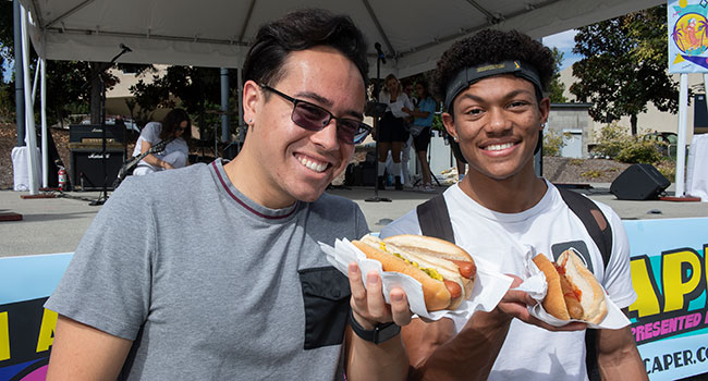 Two male students pose for a photo while holding Hot Dogs.