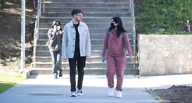 Two students walking across campus. One person is wearing a mask.