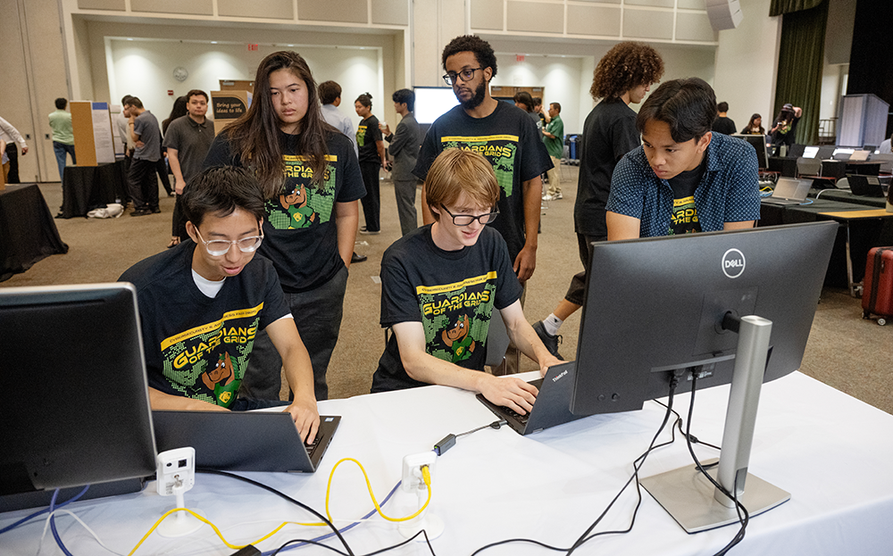 A group of students participate in the cybersecurity fair with their laptops wearing shirts that say Guardians of the Grid.