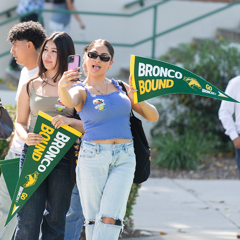 A female incoming student takes a selfie while holding a flag that says Bronco Bound 