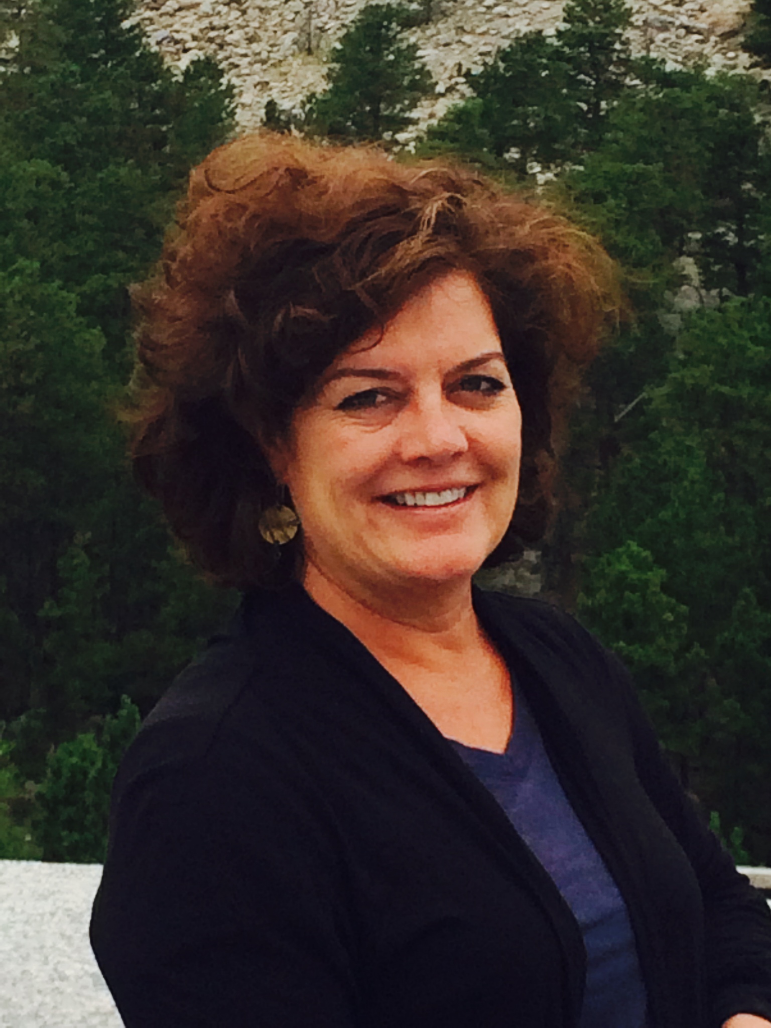 Photo of Jeannie Denholm who is a lady with short curly hair. She is smiling and is wearing a black sweater with a navy blue v-neck shirt under. She is shown in front of green bushes.