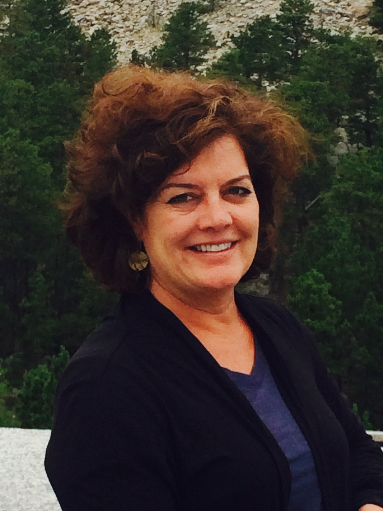 Photo of Jeannie Denholm who is a lady with short curly hair. She is smiling and is wearing a black sweater with a navy blue v-neck shirt under. She is shown in front of green bushes. 