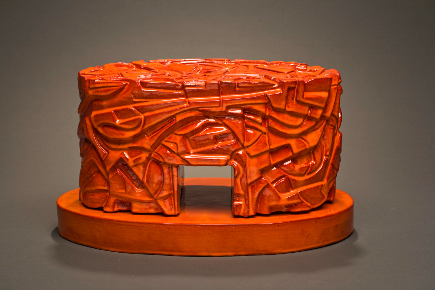 Francisco Jimenez's ceramic sculpture. It is bright red, and very glossy. The sculpture is shaped similar to a cake, but has a tiny square tunnel going through it. The sculpture is on a red disk.