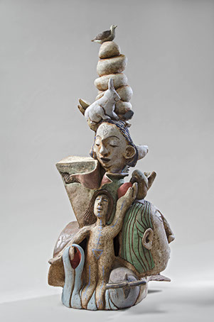 ceramic sculpture of a woman's head, another head on the side, a twist on the top of the woman's head with a bird sitting on top, a rabbit sitting on the woman's head, and a small nude person standing at the bottom of the sculpture 
