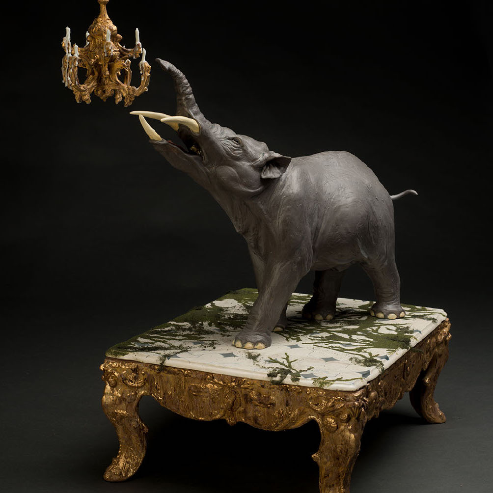 Ariel Bowman, Passe Dore, 2017. Ceramic, flocking, gold leaf, wax. 36 x 22 x 14" Courtesy of the artist. A large elephant like animal with three tusks pointing its head towards a chandelier while standing on a table with gold legs