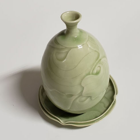 Nora Chen, The Mystic Leaf Bottle and Saucer, 2018. Wheel-thrown, hand-carved, high-fired reduction 6 x 4.5 x 4.5” Courtesy of the artist. A round spherical bottle on top of a saucer. Objects have a light green color and a leaf design placed around