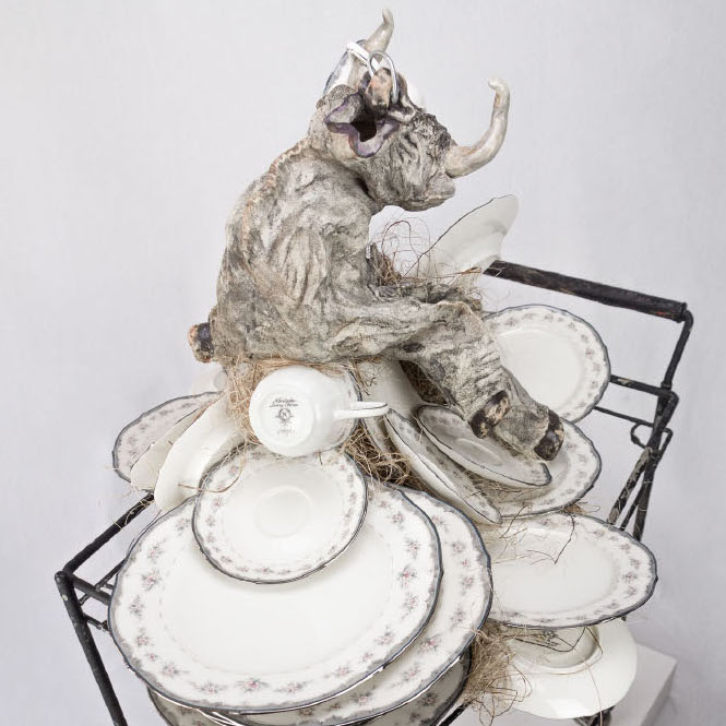 Gina M., Handle with Care, 2017. Assemblage floorpiece: ceramic, artist china, vintage found objects, coco mat. 46 x 32 x 47” Courtesy of the artist. The ‘bull in a china shop’ idiom is turned on itself in the ceramic and oxide sculpture Handle with Care. A toy bull, after assaulting a stack of china, twists with rage as a shattered platter pierces his side and stuffing spills out. I used my own wedding china to create this piece giving the china a chance to get even  .  In all my work there is a whimsy with a dark side. My personal narrative uses innocent childhood imagery like teddy bears, toys, and puppets to create the reactionary expressions of my inner emotional life. When something happens to me and triggers a buried emotion, a lost sentiment or a hidden pain, I must reconstruct and resurrect it outside of myself and find the story behind it.     Combining assemblage with ceramics fills my current body of work. The homespun construction and textured surfaces simulate threadbare fabric and tattered fur.     I select materials based on their authenticity to my process. I choose clay because of its fragility, its relationship to the earth, and its tradition in arts and craft. I incorporate recycled materials such as wood and found objects because of their nostalgia and reference to aging, decay and decomposition.