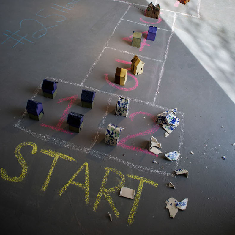 Matt Rose, Moving: A Life in 25 Houses,  2019. Ceramic and chalk floorpiece installation, composed of several ceramic parts/objects. 7 x 4 x 4” each house. Courtesy of the artist. A series of small house sculptures places on top of a chalk hop-scotch drawing