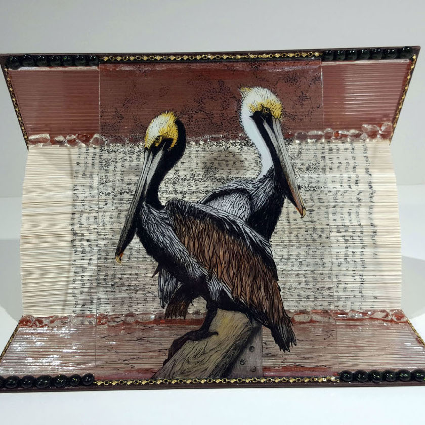 Nanci Schrieber-Smith, The Pelican Brief, 2019. Mixed media assemblage 7 x 9.8 x 7” Courtesy of the artist. Two pelicans  painted on glass in front of a book.