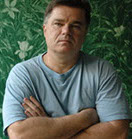 Photo of Michel David. He is a fair complected man in a light blue t-shirt. He has his arms crossed and is shown in front of a green wallpapered wall.