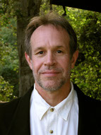 Photo of Stanton Hunter, a man smiling while wearing a black and white suit. He has a beard and is standing in front of trees.