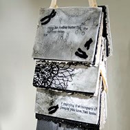 Gretchen Schermerhorn, "If Enjoying" dangling from a thick light brown thread, hangs three book-like-objects. Coming from the book there are what resembles paper feathers.