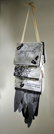 On a thick ribbon, three book-like- objects hang from it. They have abstract illustrations in black, white, and grey. At the bottom there is paper coming out that resemble the shapes of feathers.