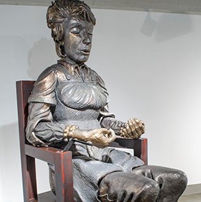 Carol Ann Klimek, "Meditator" sitting in a large chair, there is a statue of a figure meditating. 