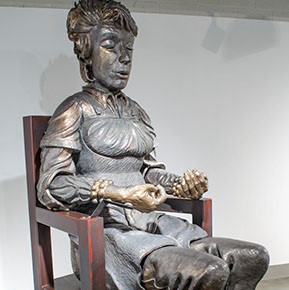 Carol Ann Klimek, "Meditator" sitting in a large chair, there is a statue of a figure meditating.