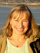 Photo of Liza Folman. She is smiling wide and has hair that is cut to her shoulders. Her dirty blond hair is also cut with front bangs. She is shown in front of the ocean.