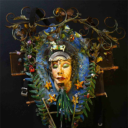 assemblage: clay with encaustic, Hol-lywood film can, typewriter keyboard and found objects sculpture of a person surrounded by green foliage (looks like a displayed wreath)