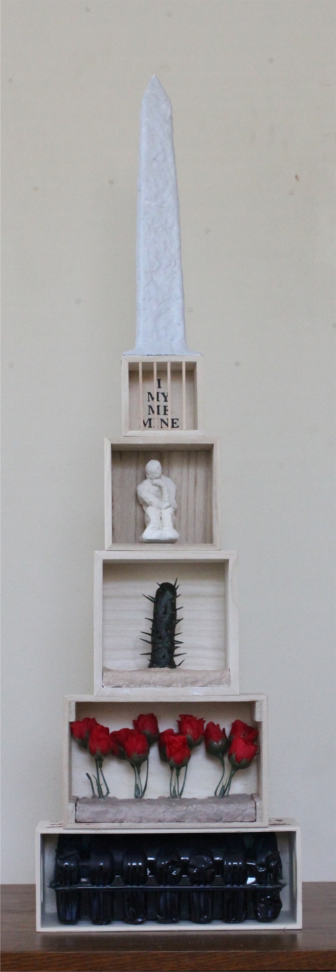 a six-leveled sculpture holding a black conatiner at the bottom, red roses above, then a single cactus, above that there is a small thinking sculpture, then there is a level that has little wooden bars with the words "I MY ME MINE" behind the bars and then the Washington Monument on top. 