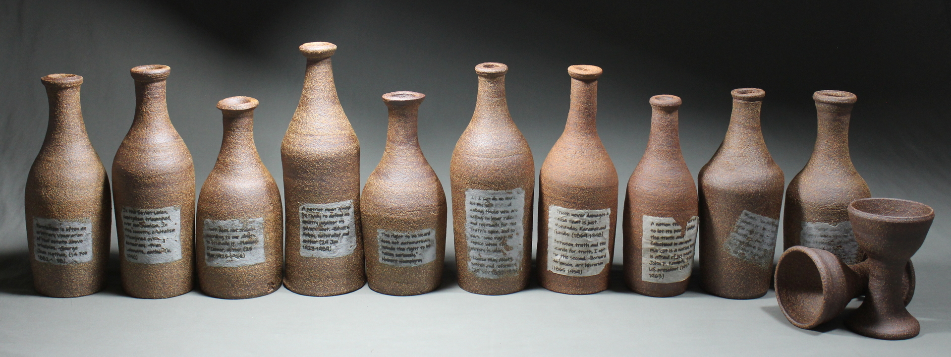 12 various sizes brown clay jugs and 2 chalices. The jugs have a piece of paper plastered on the front of them with a short blurb of text on it.