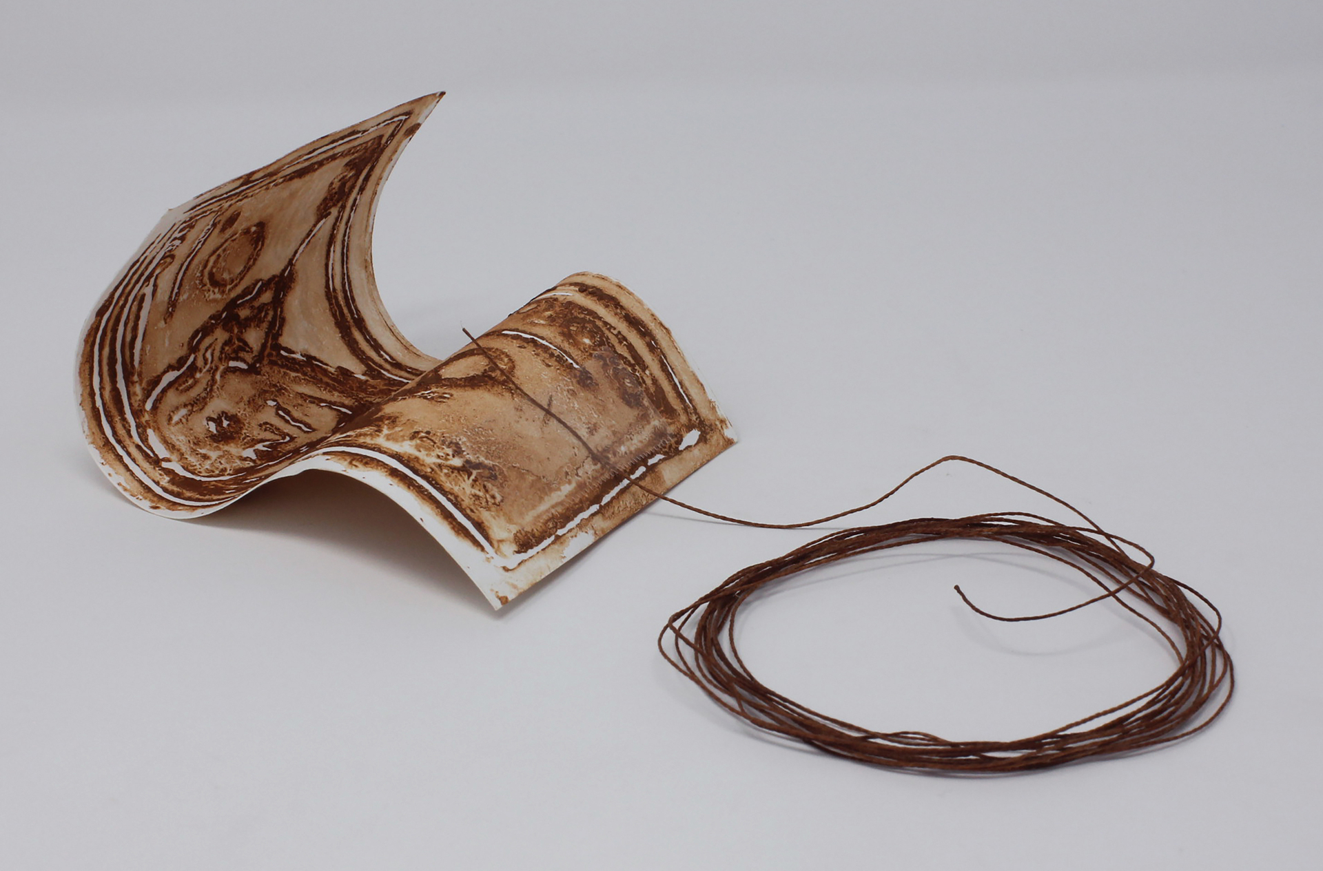 brown, rust-textured thin clay sculpture of a dollar will with a dark brown string taped to it. The string is laying in a circle right next to the dollar bill which is curled. 