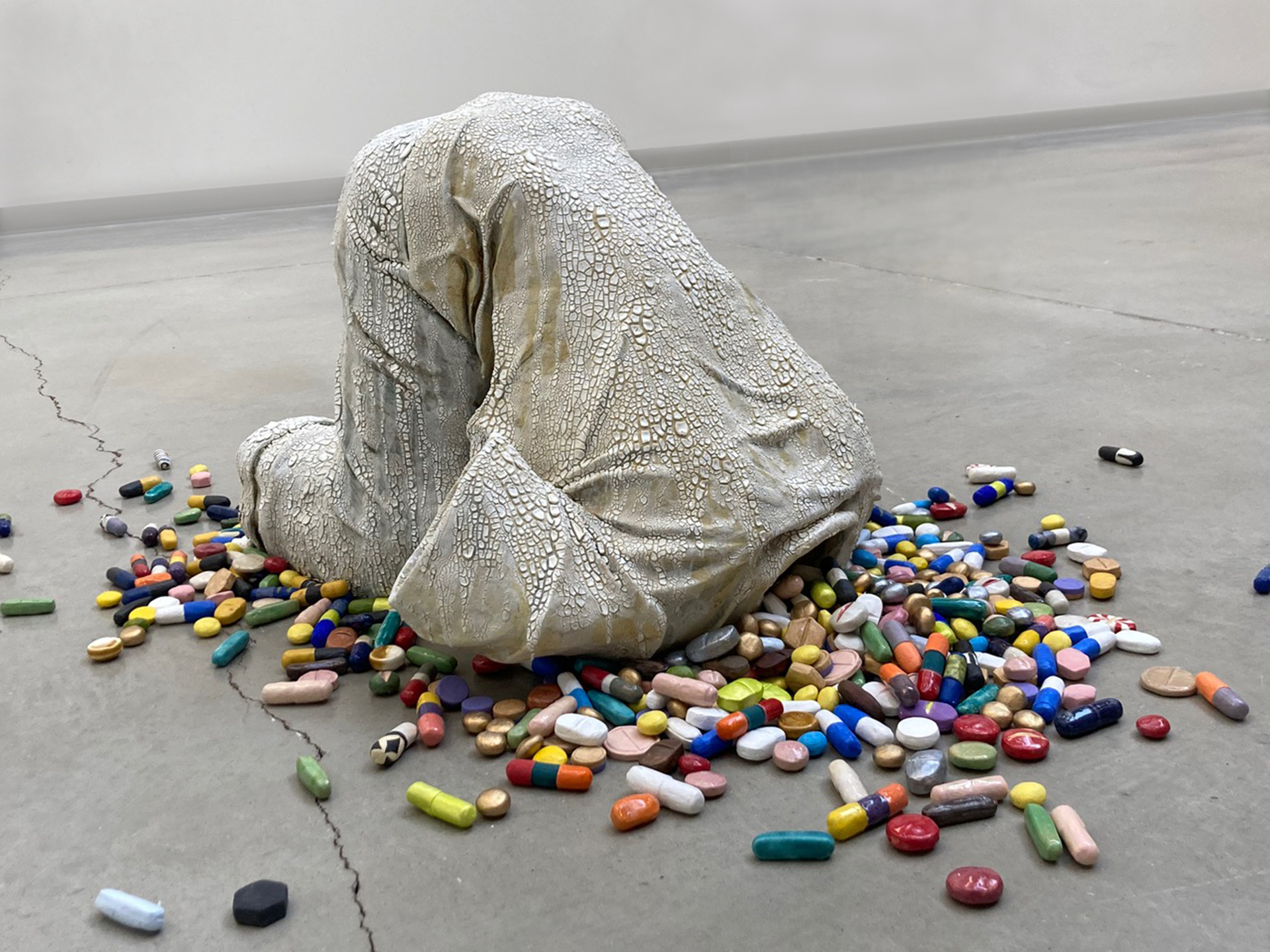 grey sculpture of a person's clothes in the shape of them. It looks like a person on their knees with their torso bent forward so their head would be on the floor except the sculpture only shows how the person's shirt and pants would look if the person were in this position. Various different color, shape and size pills spill out of the neck of the shirt where the person's head would be.