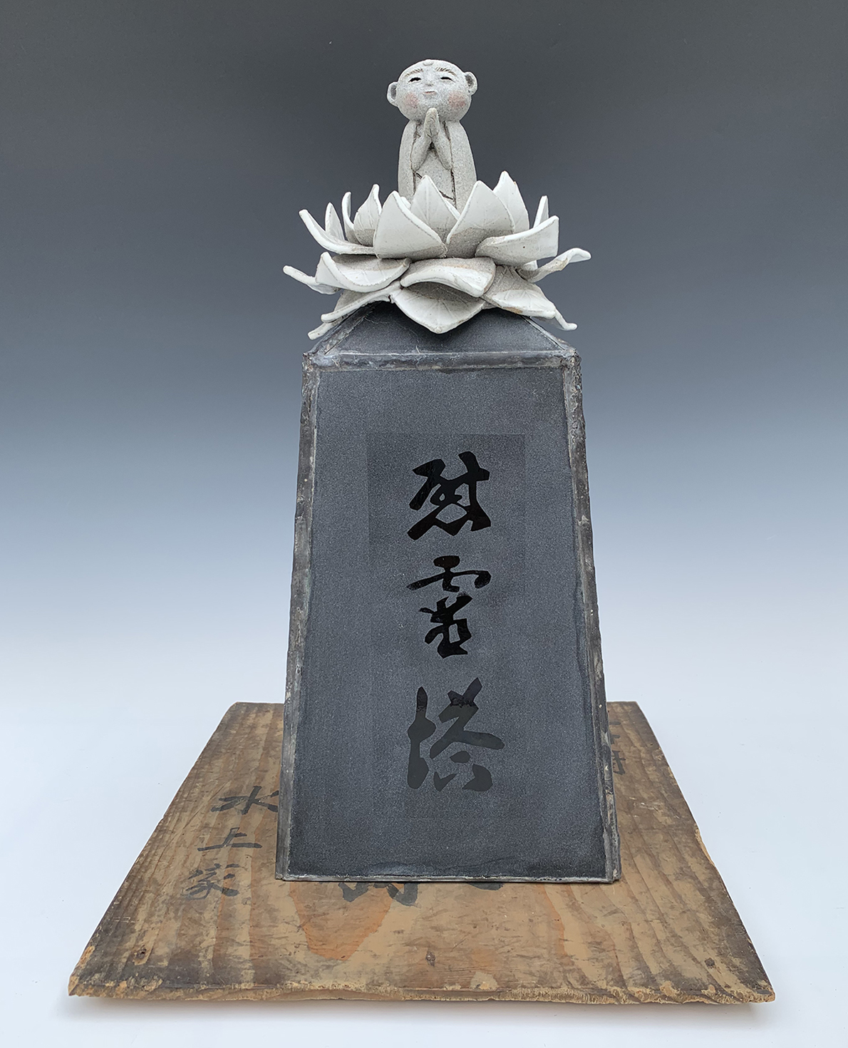 sculpture of a dark grey obelisk with 3 calligraphic asian characters written in a collumn. There is a light grey lotus flower atop the obelisk and a small buddah figure with pink cheecks and hands raised into a prayer position sitting in the lotus flower. The sculpture sits on a worn piece of wood.