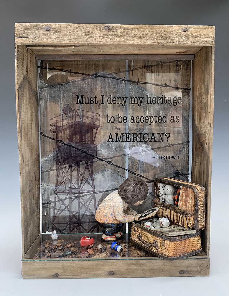 sculpture in a wooden box that has an open side. In the wooden box there is a clay woman leaning over a suitcase, examining what looks to be a photograph. There is a quote behind her that says 'must I deny my heritage to be accepted as an AMERICAN? -Unknown