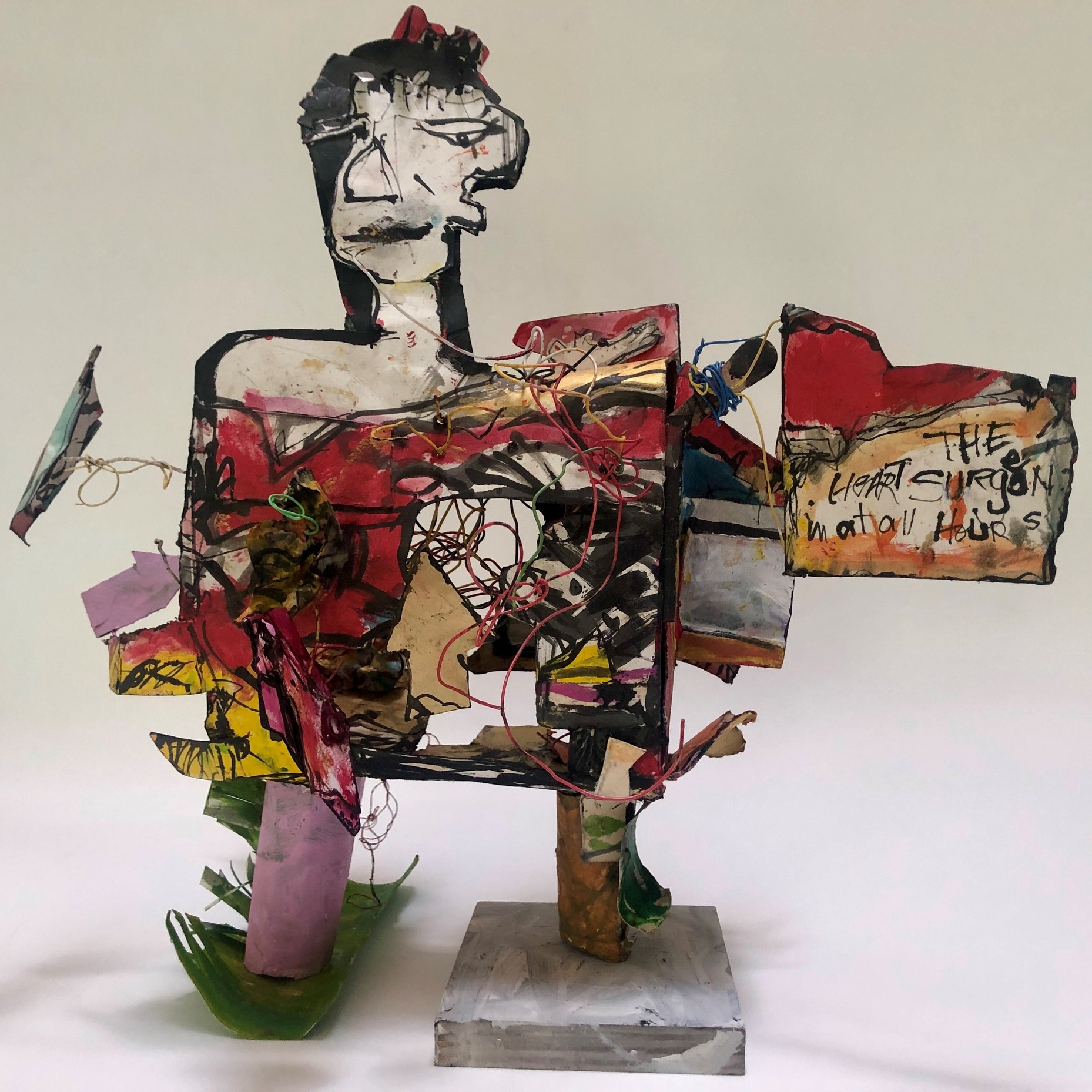 an abstract sculpture with a face, square body, and two feet standing on two different types of materials with a sign with the saying "The Heat Surgeon in at all Hours".