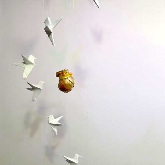 a suspended golden human heat with white paper-like cranes suspended around the heart. 