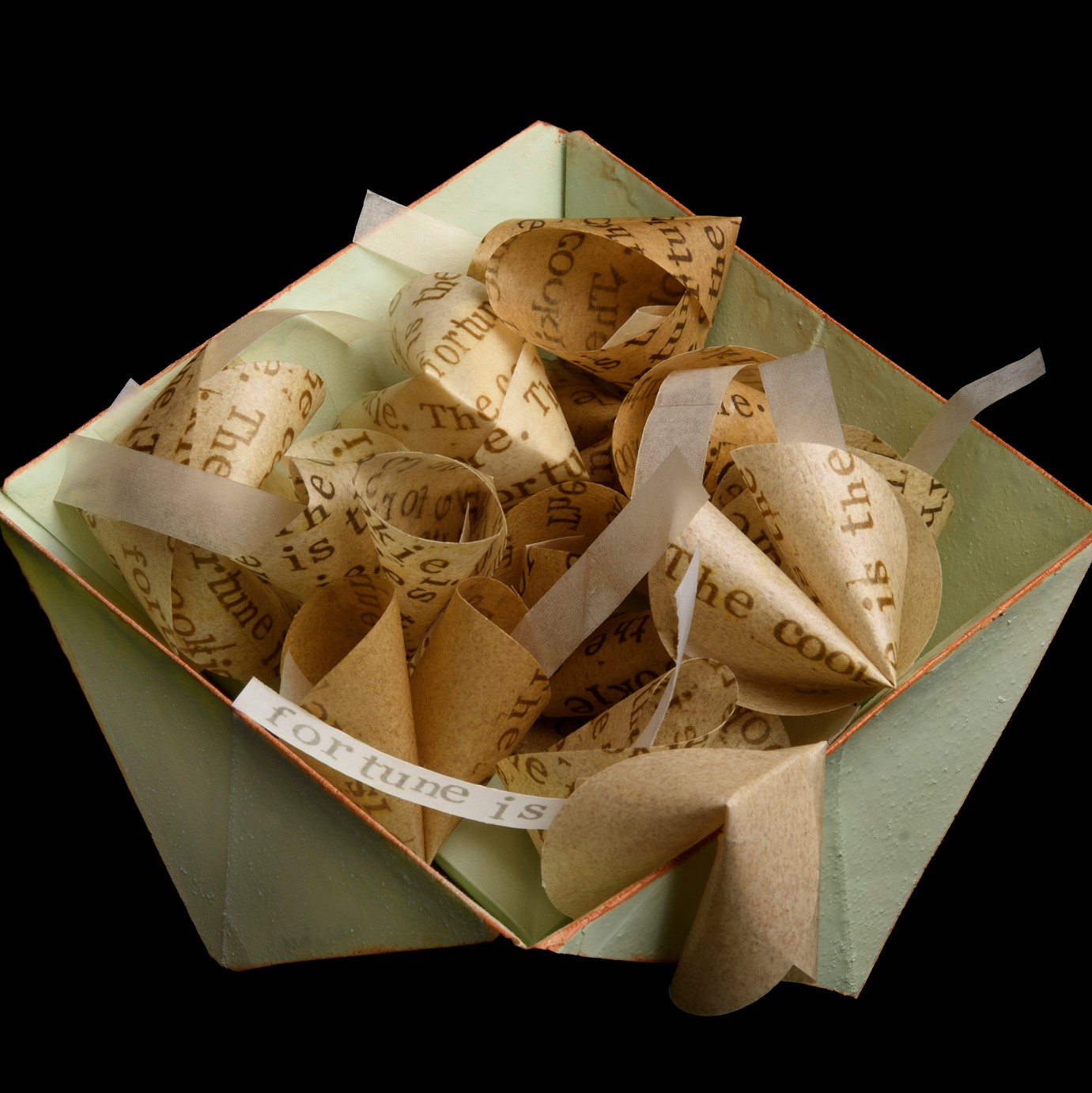 a mint green, paper oragami bowl holds multiple fortune cookies with handpritned messages on them that say, "The fortune is the cookie. The cookie is the fortune," on both the fortune cookie and the paper inside. 