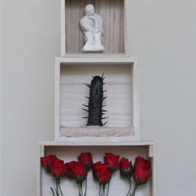 a six-leveled sculpture holding a black conatiner at the bottom, red roses above, then a single cactus, above that there is a small thinking sculpture, then there is a level that has little wooden bars with the words "I MY ME MINE" behind the bars and then the Washington Monument on top. 