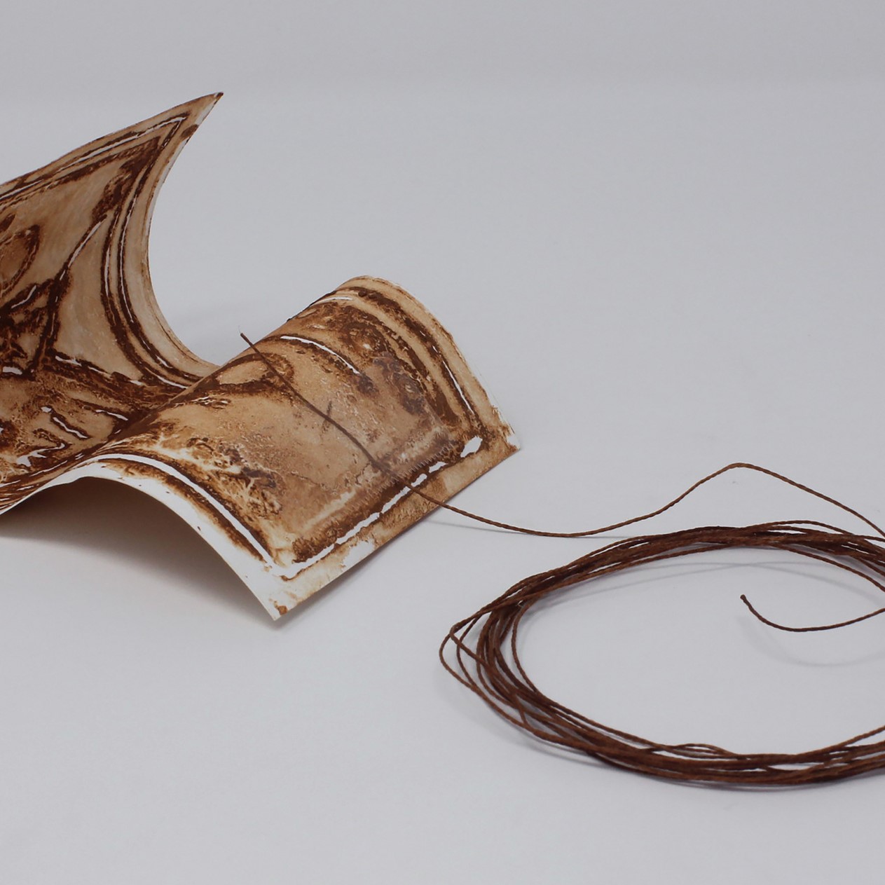 brown, rust-textured thin clay sculpture of a dollar will with a dark brown string taped to it. The string is laying in a circle right next to the dollar bill which is curled. 