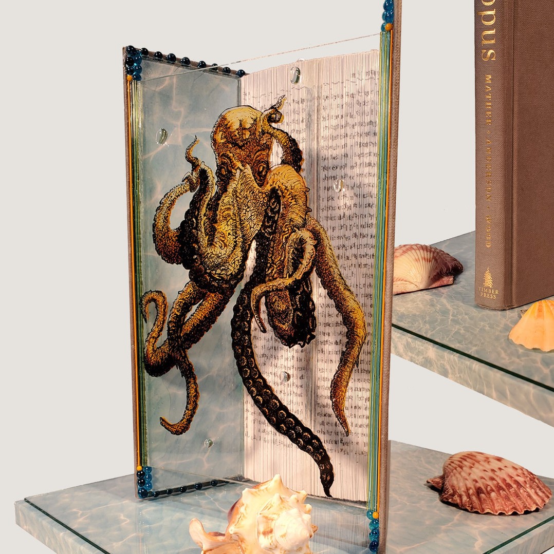 book with brown cover and spine that says 'Octopus' in gold. The book is open and the pages are folded in. A clear piece of acrylic or plexi has an illustration of an octopus on it and is placed between the front and back of the book propping it open. The book and plexi are sitting on a rectangular base with an image or painting that resembles the surface of water. There are various seashells around the sculpture.