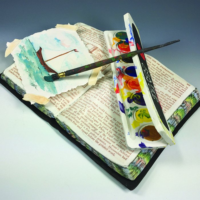 Open book with a colorful fore-edge. A watercolor set with a painbrush and a painting of a sailbot rest on top of the open book. This is all made of clay.