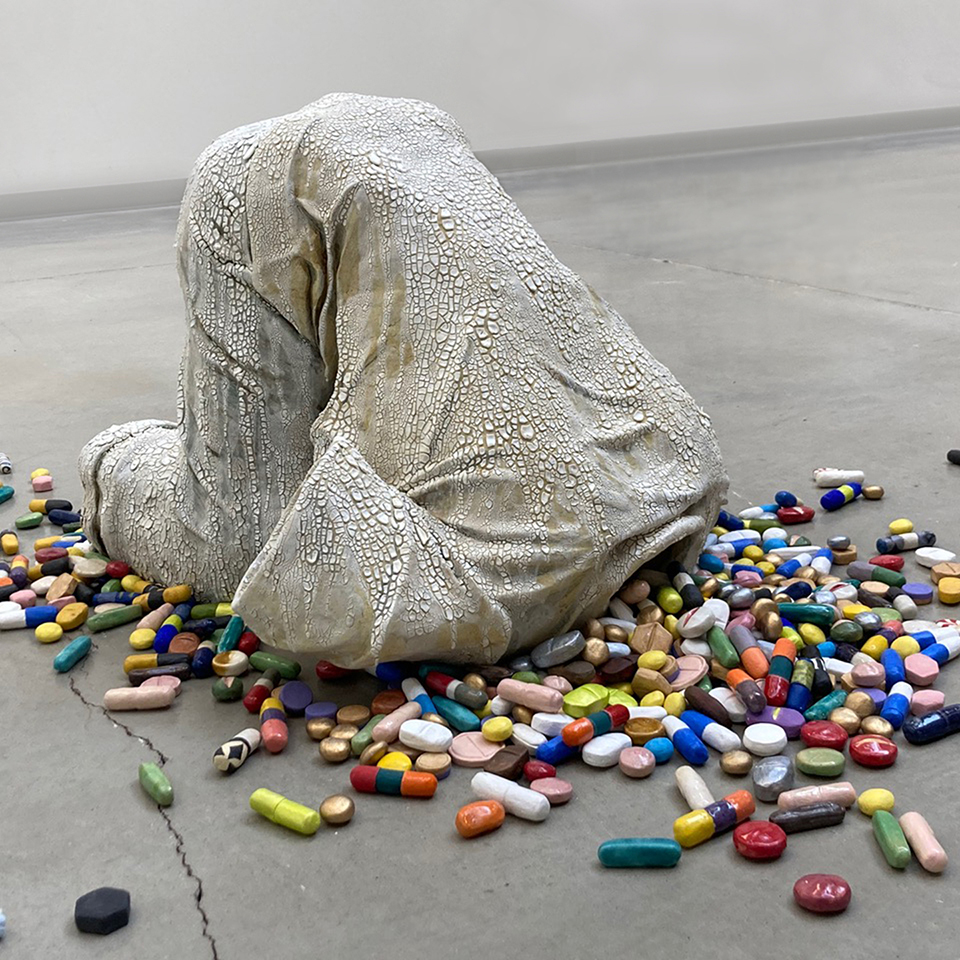 grey sculpture of a person's clothes in the shape of them. It looks like a person on their knees with their torso bent forward so their head would be on the floor except the sculpture only shows how the person's shirt and pants would look if the person were in this position. Various different color, shape and size pills spill out of the neck of the shirt where the person's head would be.