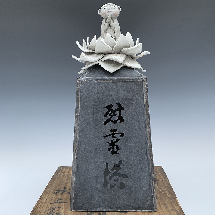 sculpture of a dark grey obelisk with 3 calligraphic asian characters written in a collumn. There is a light grey lotus flower atop the obelisk and a small buddah figure with pink cheecks and hands raised into a prayer position sitting in the lotus flower. The sculpture sits on a worn piece of wood.