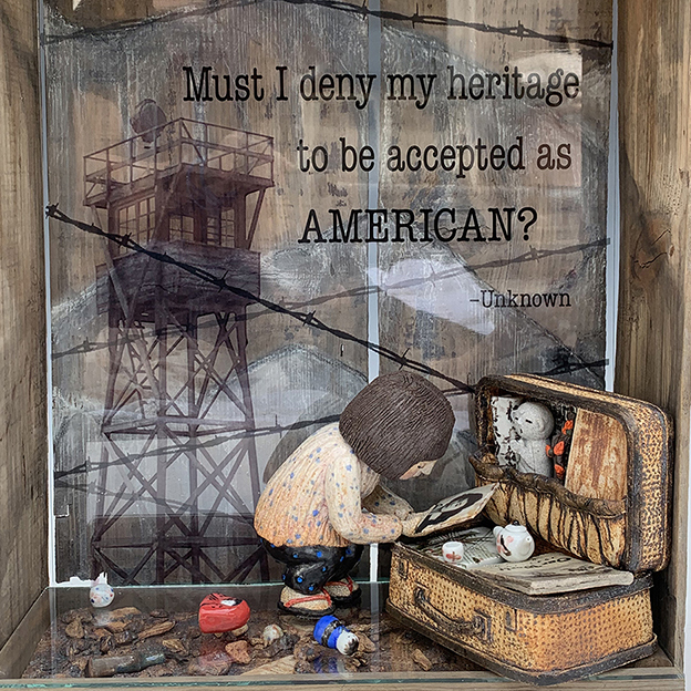 sculpture in a wooden box that has an open side. In the wooden box there is a clay woman leaning over a suitcase, examining what looks to be a photograph. There is a quote behind her that says 'must I deny my heritage to be accepted as an AMERICAN? -Unknown