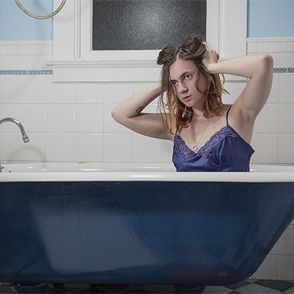 Self portrait photograph of a person in a blue dress with both hands on their head in their hair, sitting in a bathtub