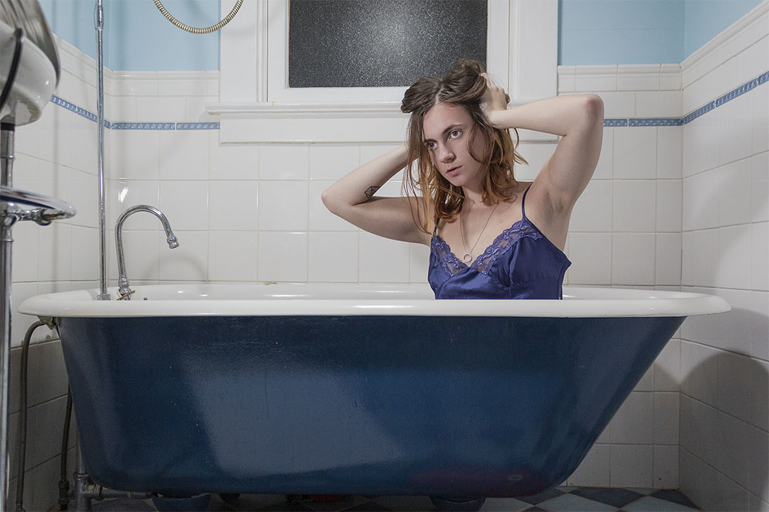 Self portrait photograph of a person in a blue dress with both hands on their head in their hair, sitting in a bathtub