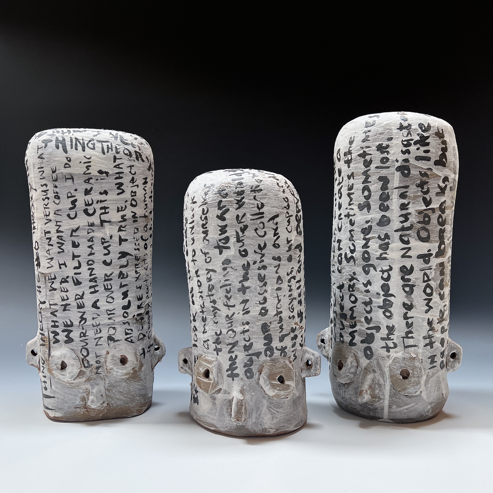  Three ceramic sculptures resembling elongated heads, each with stylized facial features, covered in handwritten black text that wraps around the forms, creating a striking contrast against the white and gray surface of the pieces.