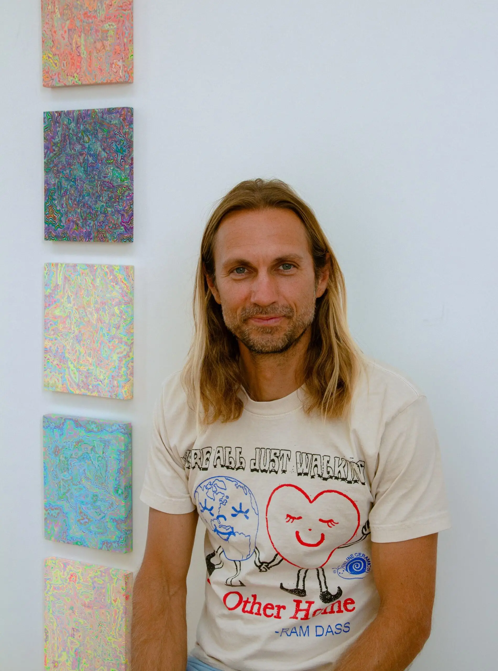 Man with long blond hair and beard, wearing a light-colored graphic T-shirt seated beside five colorful abstract paintings on a white wall.